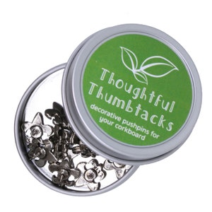 Może przedstawiać: Srebrny pojemnik z zieloną pokrywką, na kt&oacute;rej widnieje napis "Thoughtful Thumbtacks decorative pushpins for your corkboard". Pojemnik jest otwarty i zawiera srebrne pinezki z kwiatowymi wzorami.