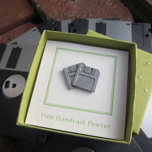 May include: A pair of small, silver-colored floppy disk pins inside a green box, presented on a white card with the text "Fine Handcast Pewter." The box rests on a background of black floppy disks and a brick surface.
