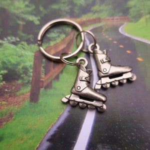 May include: A silver key chain with two roller skate charms. The charms are detailed with wheels and straps. The key chain is on a background of a winding road.