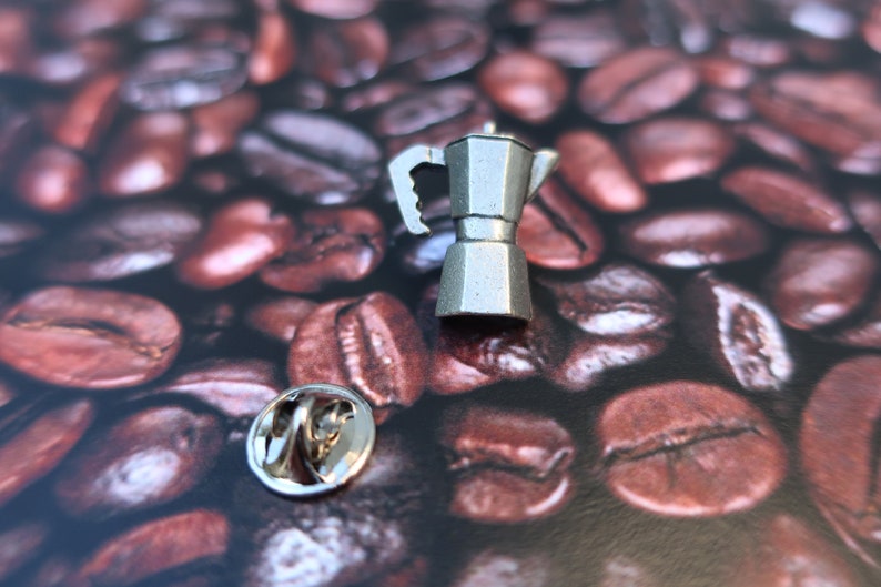 May include: A silver metal pin shaped like a traditional Italian coffee maker, or Moka pot, on a background of coffee beans.