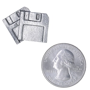 May include: Two silver-colored floppy disk-shaped objects are displayed next to a quarter. The quarter shows a profile of George Washington and the words "Quarter Dollar". The floppy disks are detailed with rectangular cutouts.
