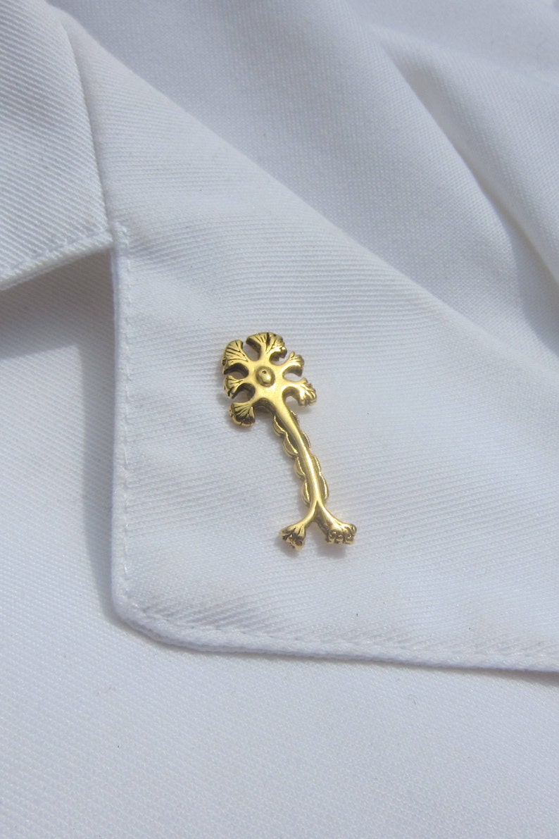 May include: A gold-colored pin shaped like a neuron, a type of nerve cell, with a detailed design. The pin is on a white fabric background.