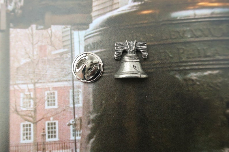 Puede incluir: Pin de plata de la Campana de la Libertad con la parte posterior redonda. El pin est&aacute; sobre un fondo marr&oacute;n con una imagen borrosa de un edificio.