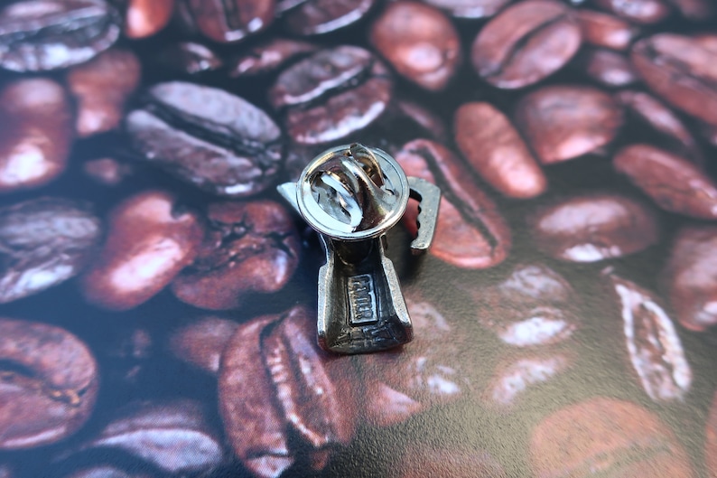 May include: Silver-toned metal pin shaped like a coffee pot with a coffee bean design. The back of the pin has a safety clasp and the word "Fender" engraved on it.