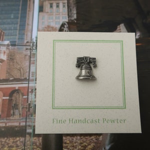 Puede incluir: Un pin de peltre de color plateado de la Campana de la Libertad, con el texto "Fine Handcast Pewter" en una tarjeta blanca con un borde verde.