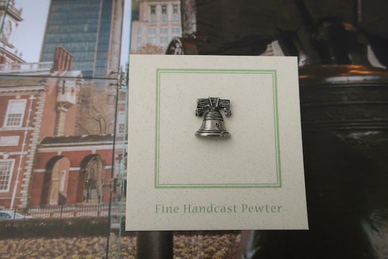 Puede incluir: Un pin de peltre de color plateado de la Campana de la Libertad, con el texto "Fine Handcast Pewter" en una tarjeta blanca con un borde verde.