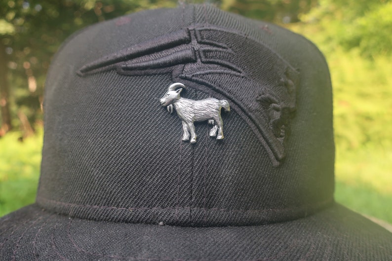 May include: A black baseball cap with a silver goat pin. The cap has a black embroidered logo on the front.