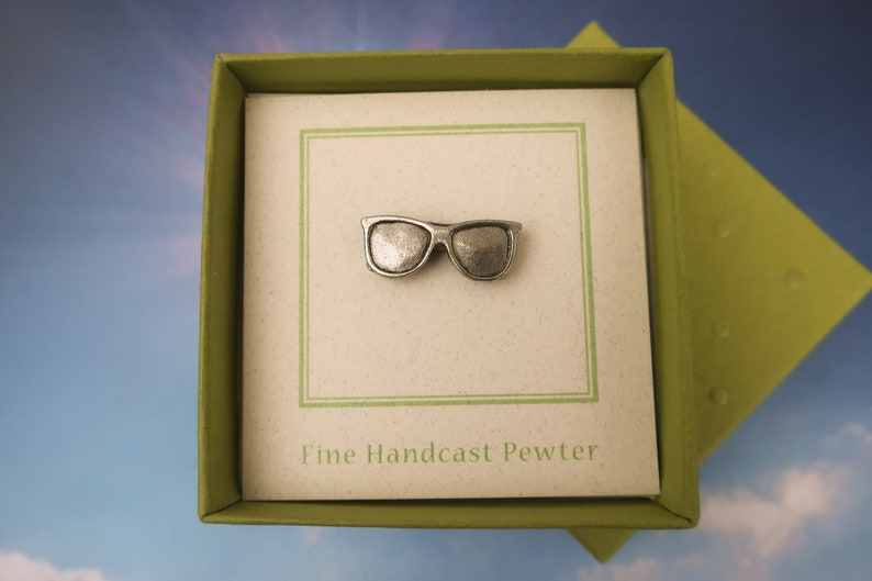 Puede incluir: Un alfiler de gafas de sol de peltre plateado en una caja verde con el texto "Fine Handcast Pewter".