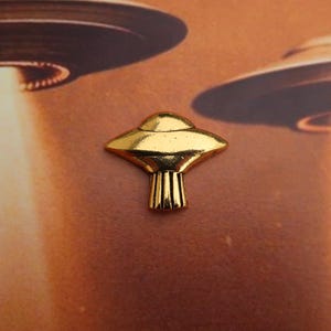 May include: A gold-colored metal pin shaped like a flying saucer. The pin is a popular collectible for fans of science fiction and UFOs.