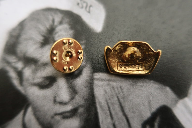 May include: Gold-toned metal pin with a raised design of a crown and the word "CREST" in all caps. The back of the pin has a circular design with a small hole in the center.