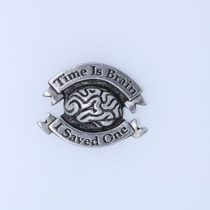 K&ouml;nnte beinhalten: Eine silberfarbene Anstecknadel mit einem Gehirndesign und dem Text "Time Is Brain I Saved One" auf wei&szlig;em Hintergrund.