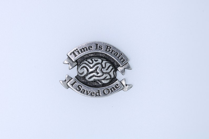 K&ouml;nnte beinhalten: Eine silberfarbene Anstecknadel mit einem Gehirndesign und dem Text "Time Is Brain I Saved One" auf wei&szlig;em Hintergrund.