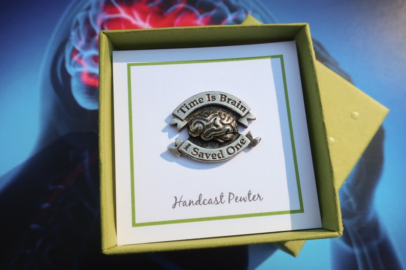 K&ouml;nnte beinhalten: Eine Zinnnadel mit einem Gehirndesign und dem Text "Time is Brain I Saved One" auf einer gr&uuml;n-wei&szlig;en Karte. Die Karte tr&auml;gt die Aufschrift "Handcast Pewter".