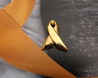 May include: A gold-colored ribbon pin in the shape of a ribbon. The ribbon is tied in a bow and has a smooth, shiny finish.