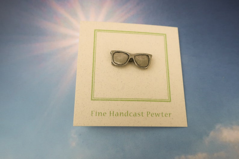 Puede incluir: Un pin de peltre plateado con forma de gafas de sol. El pin est&aacute; en una tarjeta blanca con ribete verde y el texto "Fine Handcast Pewter".