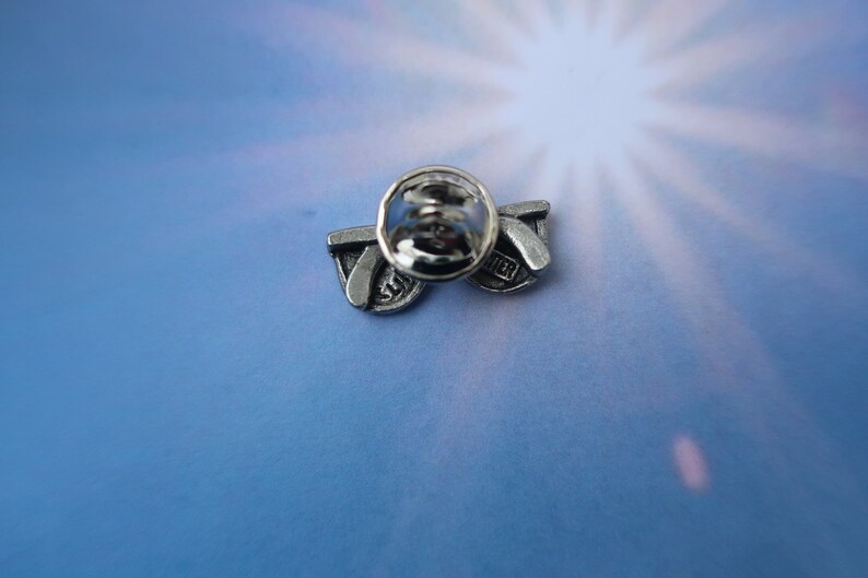 Puede incluir: Pin de tono plateado con un par de gafas de sol con la palabra "Summer" en la lente. El pin tiene un cierre de seguridad en la parte posterior.