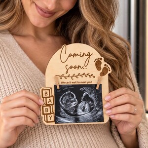 Può includere: Una targa in legno chiaro con una parte superiore ad arco che presenta il testo inciso "Coming soon.." e "We can't wait to meet you!" insieme a icone di impronte di neonato e una pila di blocchi che compongono la scritta "BABY" che sostiene un'ecografia in bianco e nero.
