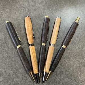 May include: Five wooden pens with gold-colored accents. The pens alternate between dark brown and light tan wood. The pens are arranged in a fan shape, with the tips pointing towards the bottom of the frame.