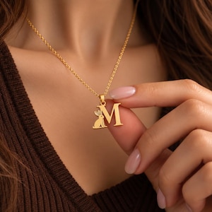 May include: A gold-toned necklace featuring a pendant shaped as the capital letter M, with a small silhouette of a rabbit sitting at the base of the letter. The pendant hangs from a delicate gold-toned chain with a classic rope-style link design.