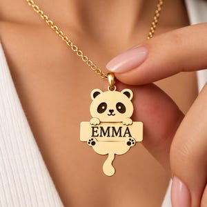 Op de afbeelding: Een goudkleurige ketting met een hanger in de vorm van een panda. De panda houdt een rechthoekige horizontale balk vast waarin de naam EMMA in zwarte hoofdletters is gegraveerd. De hanger hangt aan een fijne goudkleurige schakelketting.