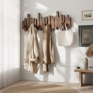 May include: A modern wooden coat rack with multiple pegs, holding a beige coat, a brown scarf, and a white tote bag. The rack is mounted on a white wall, with a round woven rug on the floor. The room is well-lit with natural light.