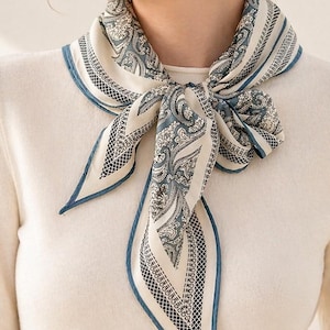 May include: A cream-colored silk scarf with a paisley pattern in shades of blue and white. The scarf is tied around the neck, with a blue border. The scarf is a fashion accessory.