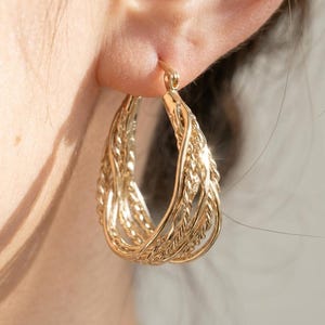 May include: A gold hoop earring featuring multiple intertwined strands with a twisted rope texture. The earring has a polished metallic finish and is secured with a hinged post mechanism that passes through the earlobe for a secure fit.
