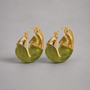 May include: A pair of hoop earrings featuring a polished gold-tone metal top section and a translucent, marbled olive green resin bottom section. The earrings have a hinged closure mechanism at the top, creating a rounded, chunky silhouette.