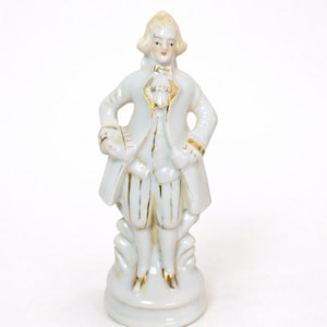 May include: A white porcelain figurine of a man in 18th-century attire. The figure is 5 inches tall and 2 inches wide, with gold accents on the clothing. The man is standing on a decorative base.
