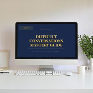 May include: A desktop computer displays a guide titled "Difficult Conversations Mastery Guide" in gold text on a navy blue background. The computer sits on a white desk with a keyboard, mouse, and a white mug. A small plant in a white vase is to the right.