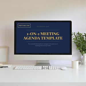 Op de afbeelding: Een computerscherm toont een donkerblauwe sjabloon met gouden tekst: "1-ON-1 MEETING AGENDA TEMPLATE." De tekst "PRINTABLE PDF" staat linksboven. Een wit toetsenbord, muis en mok staan op een witte tafel.
