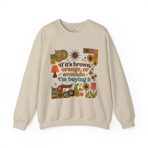 May include: A cream-colored sweatshirt with a retro design. The graphic includes an owl, sun, flowers, and fruit, along with the text "If it's brown, orange, or avocado - I'm buying it."