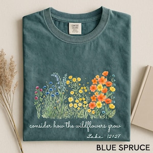 Puede incluir: Una camiseta azul verdosa con un gráfico estilo acuarela de varias flores silvestres en tonos azules, amarillos y naranjas. Debajo de las flores, el texto dice "consider how the wildflowers grow" seguido de "Luke 12:27" en letra cursiva blanca.