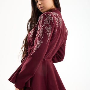 Könnte beinhalten: Burgunderfarbene formelle Jacke mit floralen Perlenverzierungen an Schultern und Ärmeln. Die Jacke hat eine Schößchen-Taille und einen passenden Gürtel. Das Outfit ist Teil einer formellen Bekleidungskollektion.