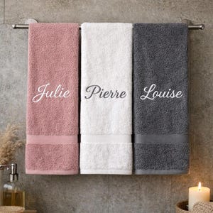 Peut inclure: Trois serviettes de bain rectangulaires suspendues à une barre métallique. La serviette de gauche est rose poudré avec le prénom Julie brodé en blanc. Celle du centre est blanche avec Pierre en gris. Celle de droite est gris anthracite avec Louise en blanc.