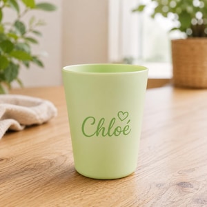 Peut inclure: Un gobelet vert clair portant le prénom Chloé écrit en lettres cursives vert foncé, accompagné d'une petite icône en forme de cœur placée au-dessus de la lettre e. Le gobelet présente une finition mate lisse et une forme cylindrique évasée.