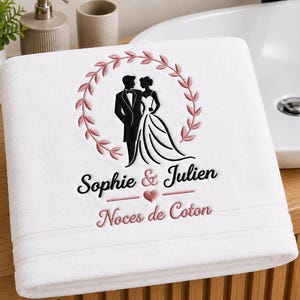 Peut inclure: Une serviette blanche avec une broderie représentant un couple en tenue de cérémonie dans une couronne de feuilles roses. Sous le couple, le texte Sophie & Julien avec un petit cœur rose, suivi du texte Noces de Coton en rose.
