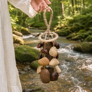 May include: A hanging mobile ornament constructed from a circular jute rope frame with a braided loop. Suspended from the frame are multiple natural elements including smooth dark brown seed pods and textured light tan dried nut shells on jute strings.
