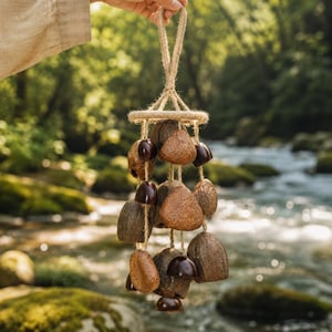 May include: A hanging wind chime with a natural, earthy aesthetic. The chime features multiple bell-shaped pieces in shades of brown and tan, suspended from a circular frame and a jute rope. The background shows a river and lush greenery.