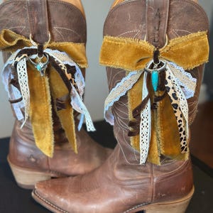 May include: A pair of brown leather cowboy boots with decorative accessories. Each boot features a mustard yellow velvet bow, lace trim, and a turquoise charm. The boots have pointed toes and a worn appearance.
