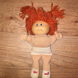 May include: A vintage Cabbage Patch Kid doll with orange yarn hair styled in two tufts. The doll has a peach-colored face, blue eyes, and a neutral expression. It wears a white diaper and cream-colored shoes with pink ribbons.