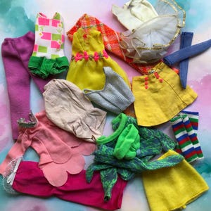 May include: A collection of doll clothing including a white and green patterned bodysuit, a yellow dress with pink trim, a yellow skirt with pink buttons, a pink top, purple trousers, a white tulle skirt with gold trim, and striped socks.