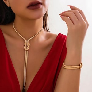 Könnte beinhalten: Goldfarbenes Halsketten- und Armband-Set. Die Halskette hat ein Knotendesign in der Mitte mit einer langen, herabhängenden Kette. Das Armband hat ein ähnliches Knotendesign. Der Schmuck wird mit einem roten Kleid getragen.