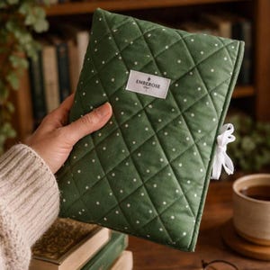 Op de afbeelding: Een groene gewatteerde boekomslag met witte stippen en een wit lint. De omslag heeft een ruitpatroon en een rechthoekig label met de tekst "EMBEROSE". De omslag wordt in een hand gehouden.