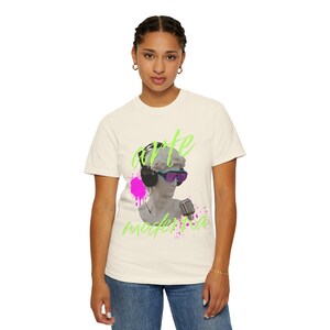 Peut inclure: T-shirt crème avec un graphisme représentant un buste portant un casque et des lunettes futuristes, tenant un microphone. Les mots "arte moderna" sont écrits en vert fluo. Une éclaboussure de peinture rose est derrière le buste.