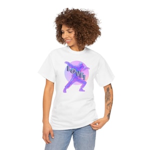 Puede incluir: Una camiseta blanca de manga corta con un gráfico de una persona con sudadera con capucha realizando un paso de baile. La figura está representada en tonos morados y azules sobre un fondo circular degradado con la palabra LOVE! en letras.