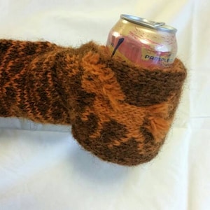 Könnte beinhalten: Ein braunes und oranges gestricktes Getränke-Cozy für eine Getränkedose. Das Cozy hat ein Muster aus braunen und orangen Streifen und ein braunes und oranges Elch-Design. Das Cozy hält eine Getränkedose mit dem Wort "Pamplemousse" auf der Dose.