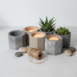 Puede incluir: Una colección de macetas y candelabros hexagonales de hormigón. Algunas macetas contienen pequeñas suculentas, mientras que otras contienen velas de té encendidas. También hay una madeja de hilo y piedras lisas.