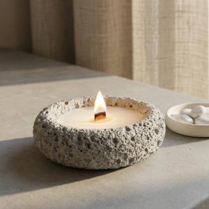 Puede incluir: Una vela encendida en un cuenco gris texturizado con forma de piedra. La vela tiene una mecha de madera y un interior de cera blanca. Un plato pequeño, blanco y redondo con caramelos blancos está a la derecha de la vela.