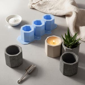 May include: A selection of hexagonal concrete planters and moulds. Includes a blue silicone mould with three cavities, a lit candle, and a small succulent plant. Also shown are a metal scoop and a bowl of white powder.
